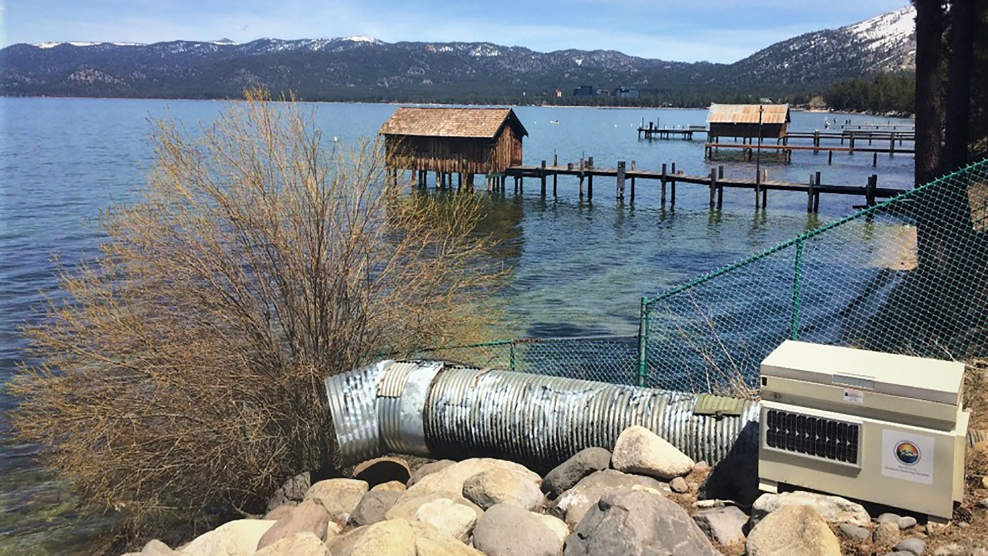 A lake with mountains, piers, boathouses, a large pipe, and some equipment on the shore.