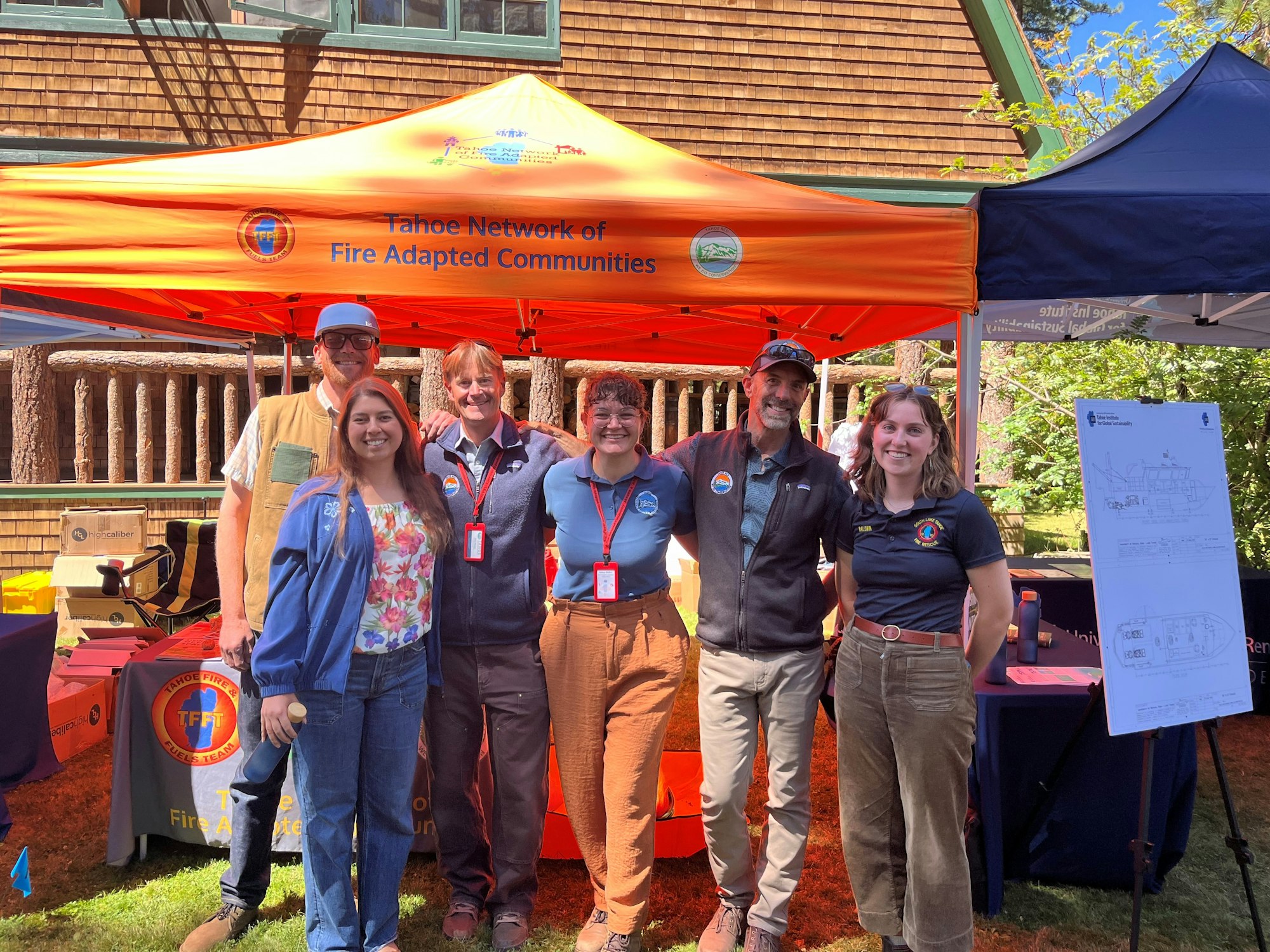 A group of six people posed under an orange "Tahoe Network of Fire Adapted Communities" tent at an outdoor event.