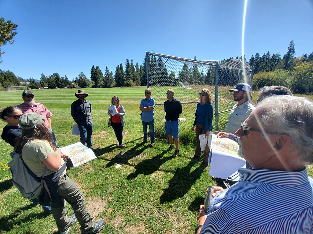 Group of people having a discussion outdoors on a sunny day, possibly during a meeting or field trip, with grass and trees around.
