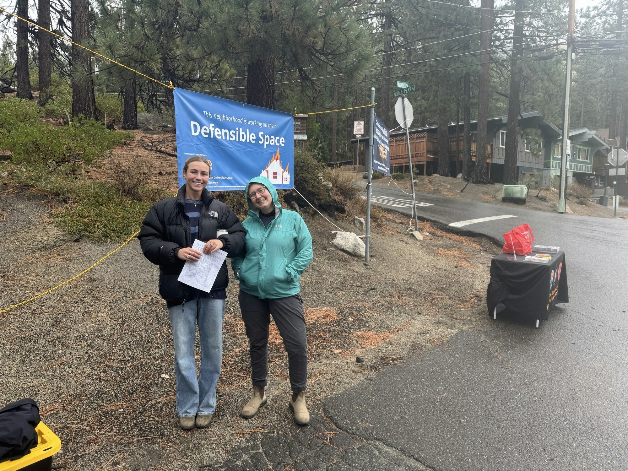 Two women stand in front of a "Defensible Space" sign in a wooded area, engaged in community safety work with a table nearby.