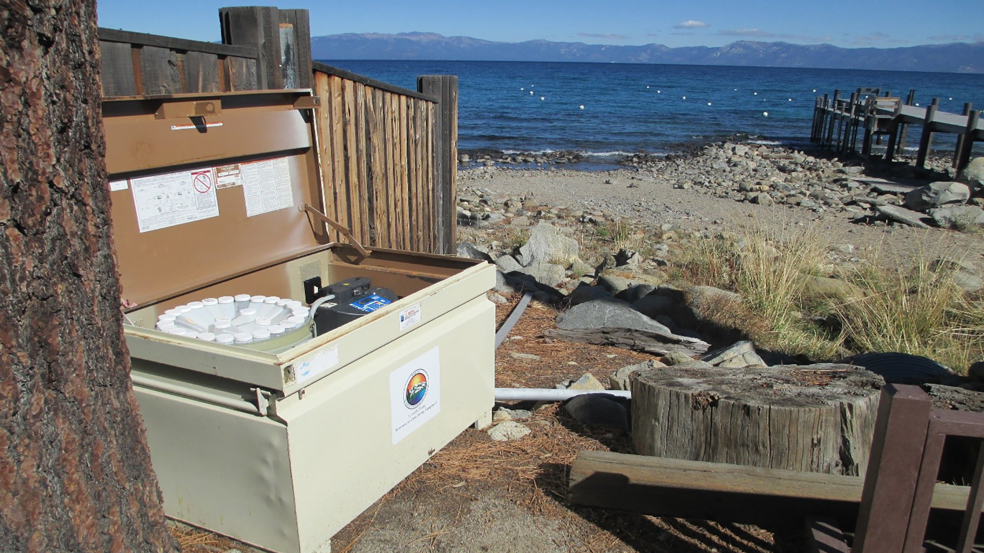 An open bear-proof container with items, by a lake with mountains in the distance.