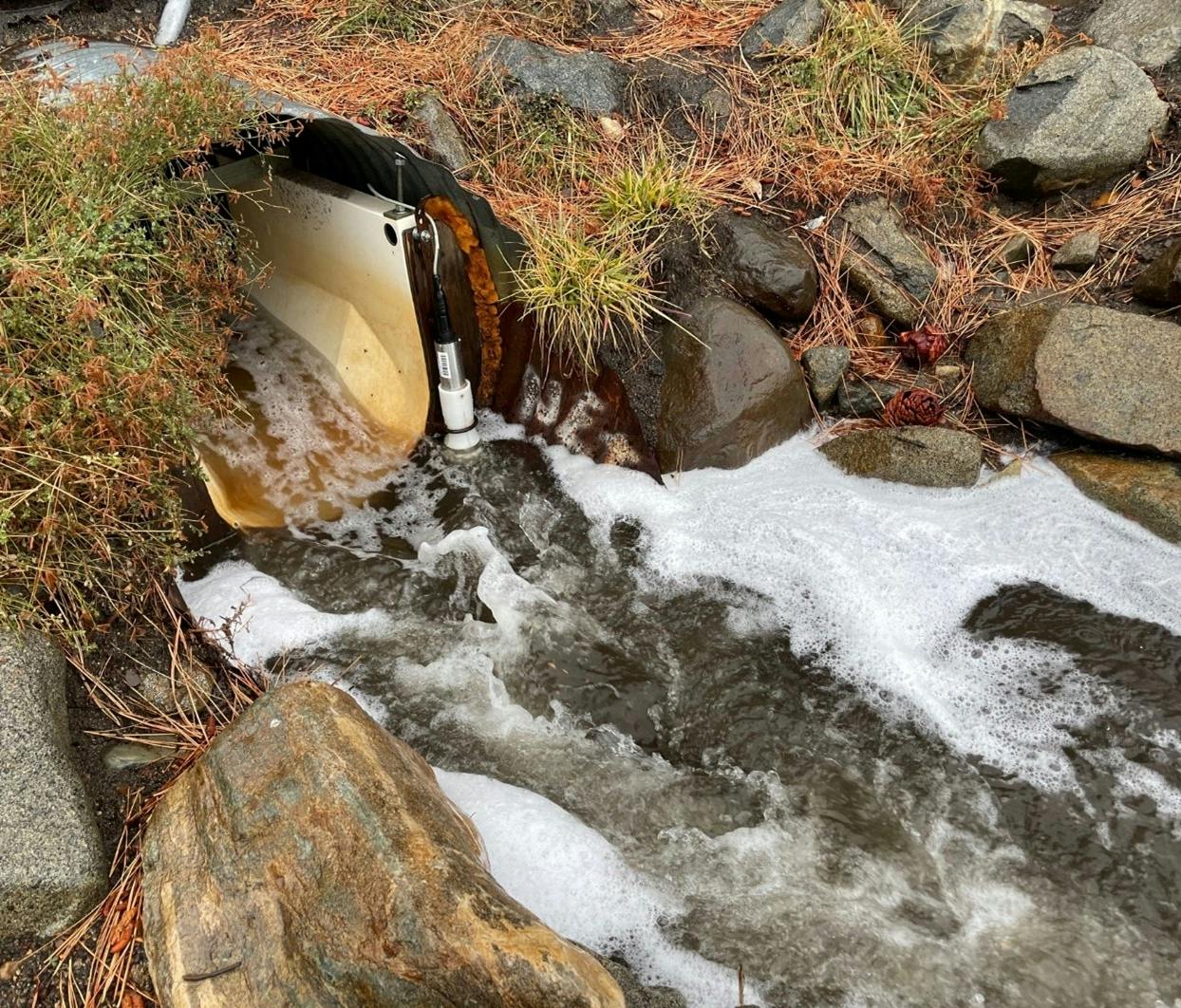 The image shows a drainage outlet with water flowing through it, surrounded by rocks and vegetation with some foam.