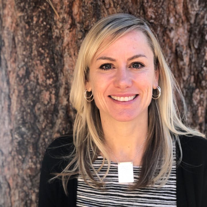 A smiling woman with a striped top, earrings, and a necklace against a tree bark background.