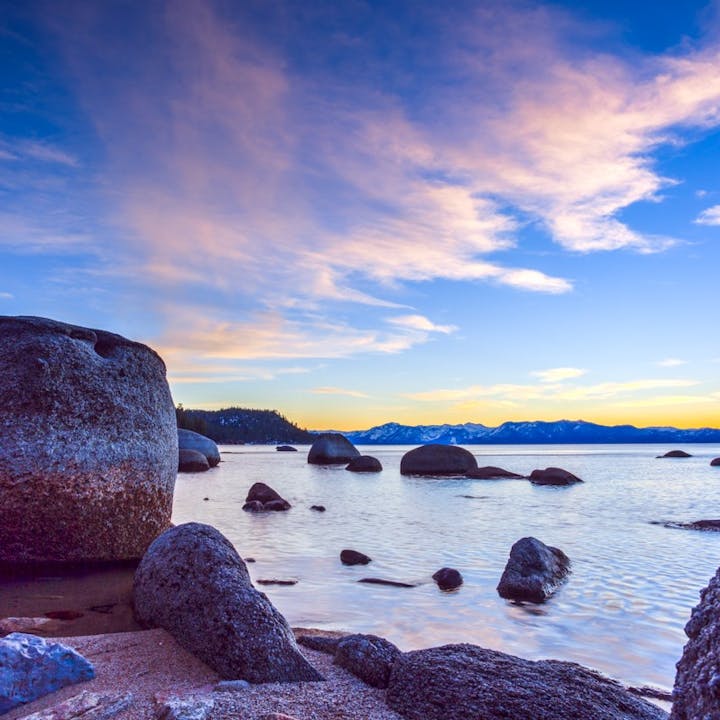 Sunset over a serene lake with scattered rocks and distant mountains under a pastel sky.