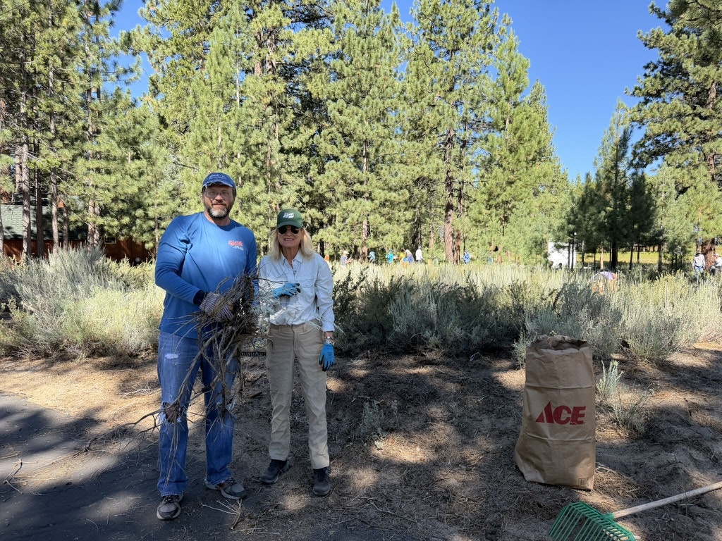 Two people are smiling while holding sticks in a green, wooded area with others working in the background.