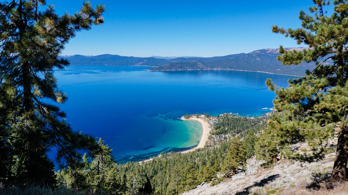 Scenic view of a clear blue lake surrounded by mountains and trees with a sandy beach in the foreground.