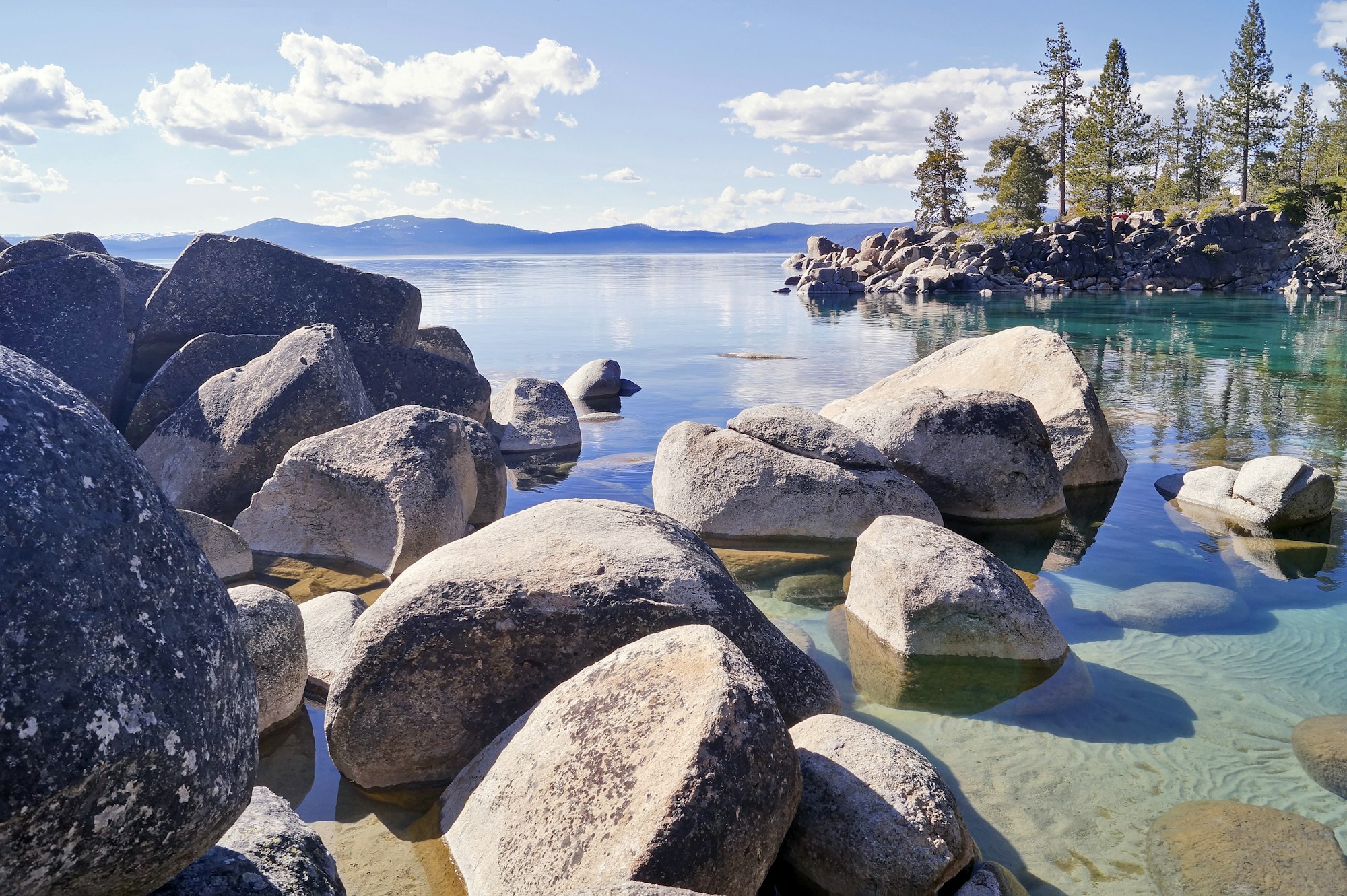 Rocks in clear water near a scenic lake with distant mountains under a blue sky.