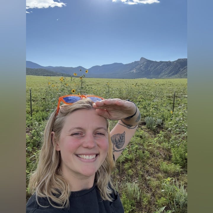 Person smiling in a field with sunflowers and mountains in the background, shielding eyes from the sun.