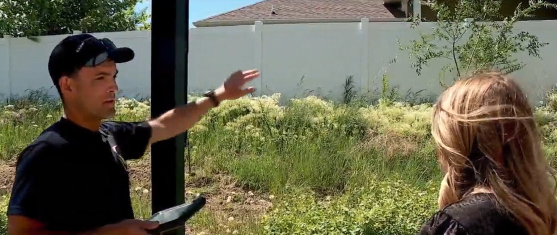 Man gesturing and talking to a woman outdoors, with lush greenery and a fence in the background.