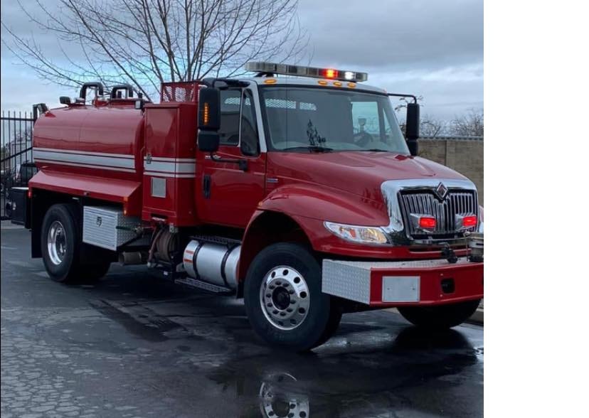 A red water tanker truck with emergency lights on a cloudy day.