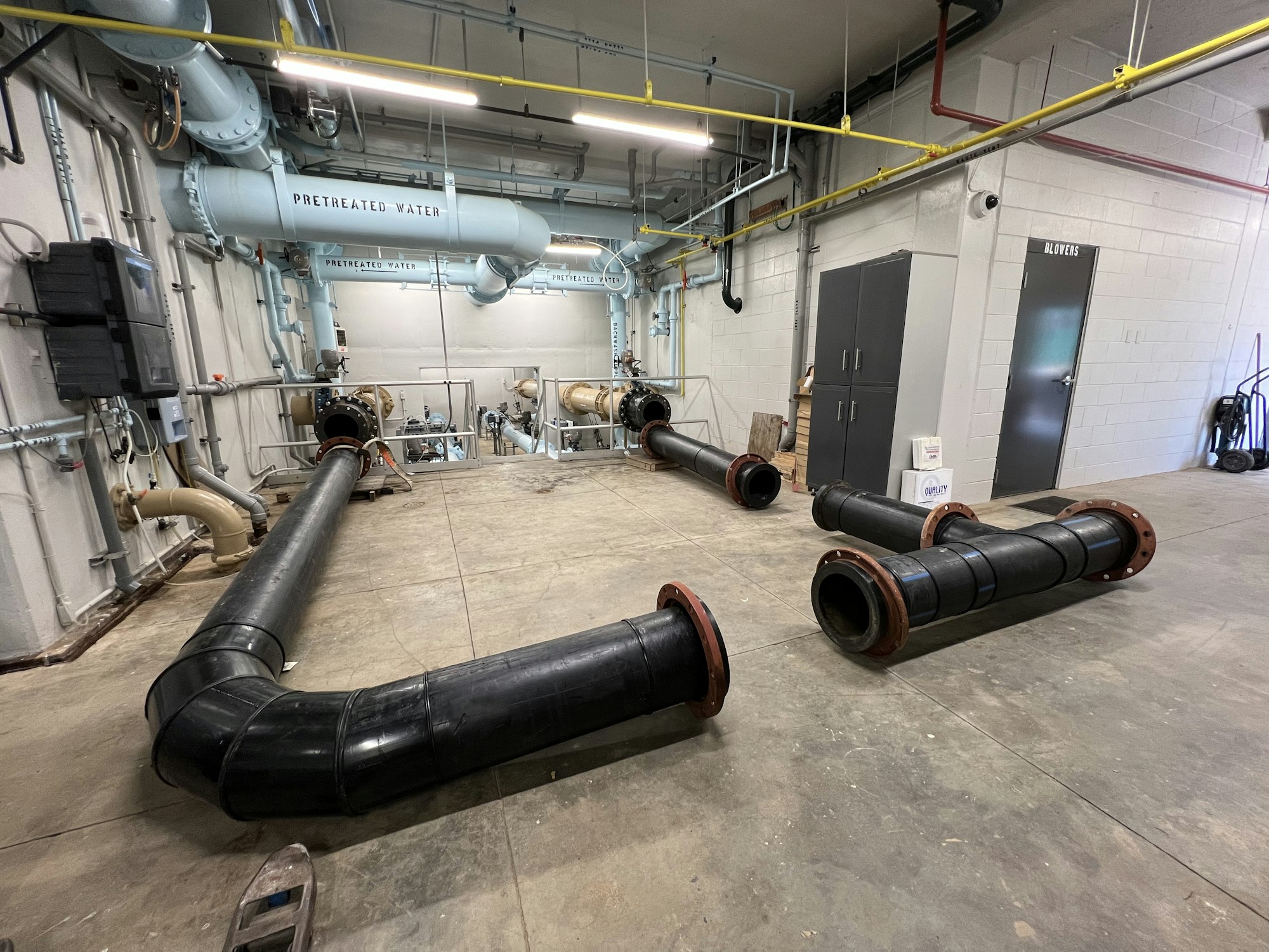 Large black piping added to water treatment plant room in order to allow construction work inside the plant. Large pipes are labeled "PRETREATED WATER.".