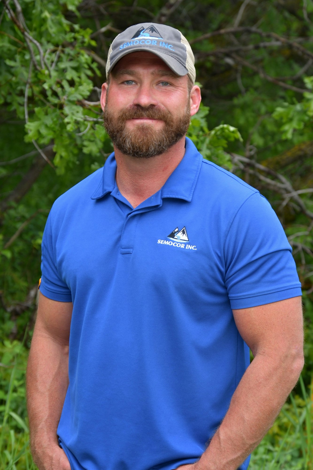 Headshot of Will in a blue half-button shirt and cap, with greenery in background.