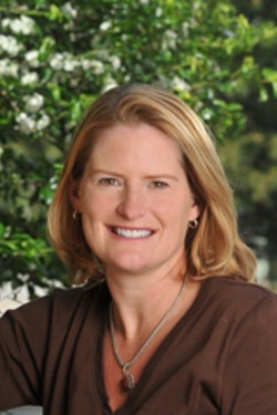 Headshot of Susan, who has short blond hair, is smiling, and is wearing a brown shirt with trees in the background.
