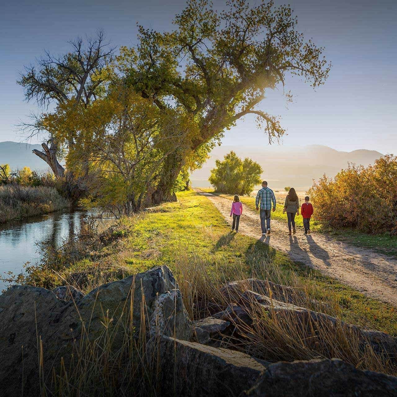 A family of four walks along a scenic path bordered by a stream and trees, under a clear sky.