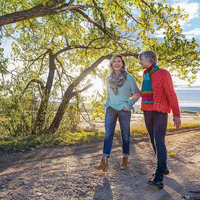 Two people walking and laughing on a sunny path surrounded by trees and greenery.