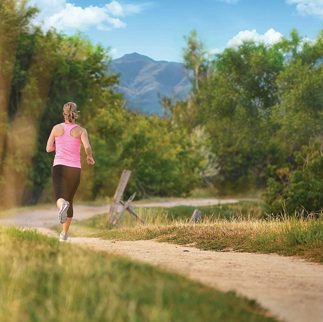 A person in a pink top runs on a dirt path through a lush, green landscape with mountains in the background.