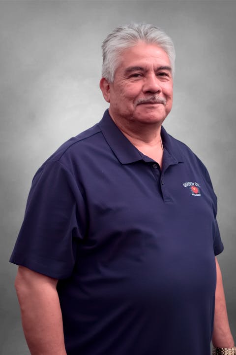 The image shows a man with gray hair and a mustache wearing a navy blue polo shirt, posing against a gray background.