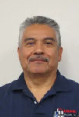 A person with short hair wearing a navy blue collared shirt, standing against a plain background.