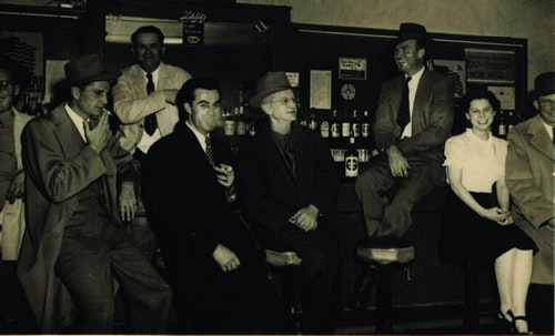 A group of people, some seated and some standing, in formal attire at a bar, with drinks and hats, in a vintage setting.