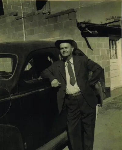 A man in a suit and hat poses beside a vintage car in front of a brick building.