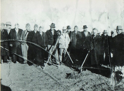 A group of men in suits and hats stand together on a dirt ground with some equipment.