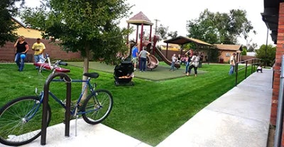 A park scene with people and children playing. There's a playground with a slide, a bicycle parked by a tree, and a stroller nearby.