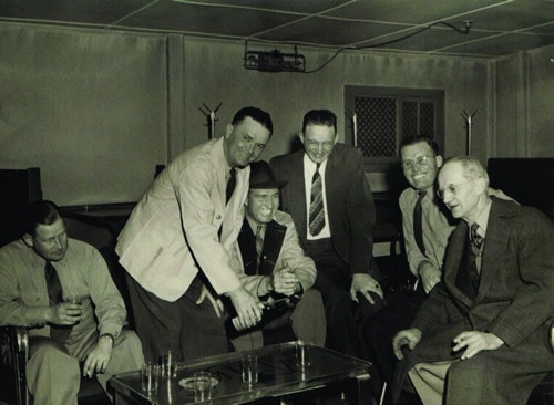 Six men in suits sitting and standing around a table, sharing drinks and smiling in a room with a low ceiling.
