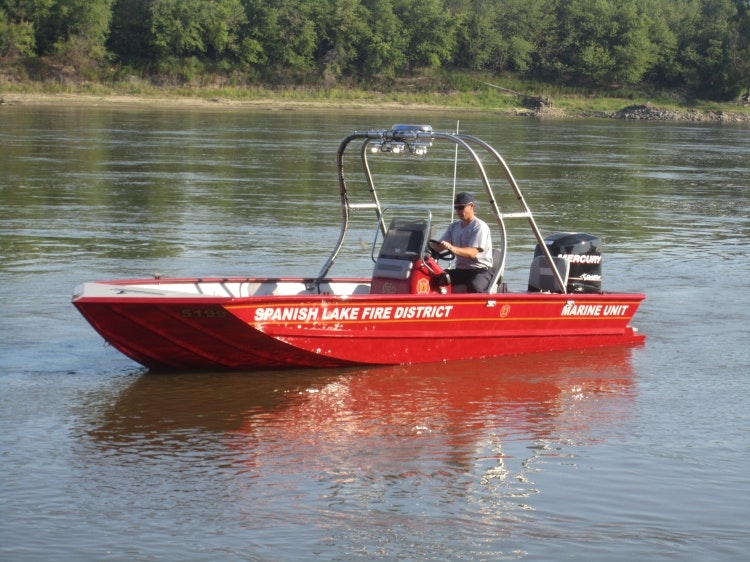 A red rescue boat labeled "Spanish Lake Fire District Marine Unit" on a river, piloted by a person.