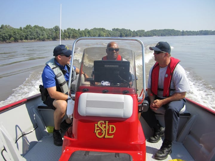 Three people in a red motorboat on a river, wearing life vests, under clear blue skies.