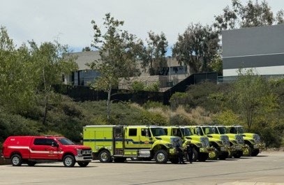 A row of yellow fire trucks and a red utility vehicle parked on the road near trees and buildings.