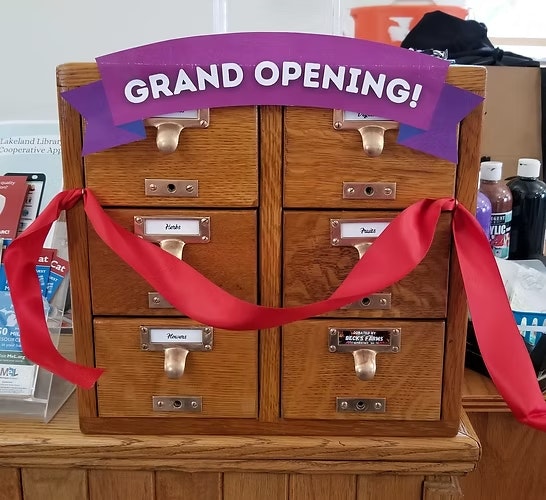 A wooden card catalog with drawers, decorated with a "Grand Opening!" sign and red ribbons, likely at a library or event.