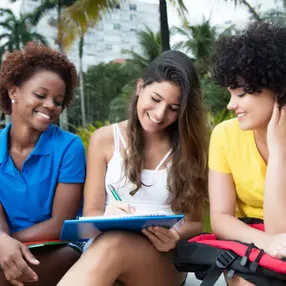 Three young women sit together outdoors, smiling and engaging, with one writing in a notebook. A vibrant, relaxed atmosphere.