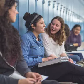 A group of students are sitting in a school hallway, engaged in conversation and smiles, with lockers in the background.