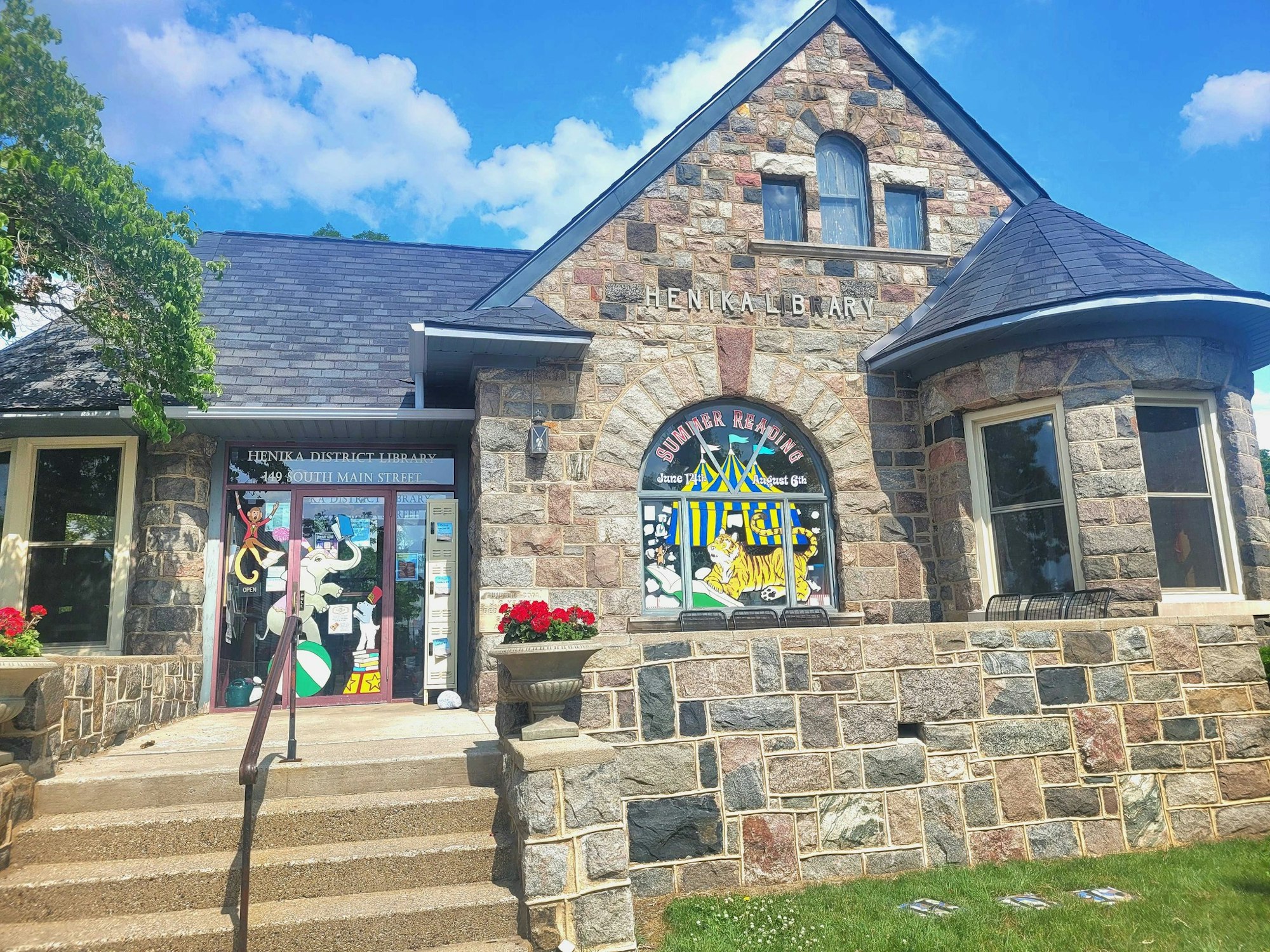 The image shows the Henika District Library, featuring a stone exterior, colorful entrance decorations, and a blue sky.