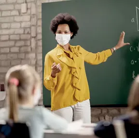 A teacher in a yellow blouse and mask instructs students in front of a chalkboard in a classroom setting.
