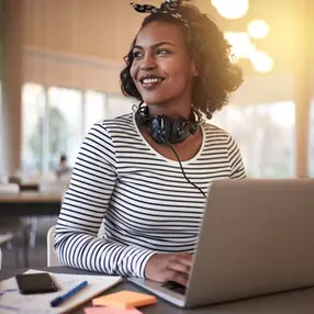 A woman with curly hair wearing headphones smiles while working on a laptop in a bright, modern setting.