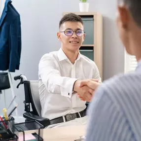 A young man in a white shirt shakes hands with someone out of view, seated at a desk in an office setting.