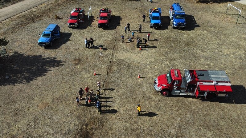 Aerial view of people and vehicles, including a fire truck, in a grassy area with activities resembling a community event or training.