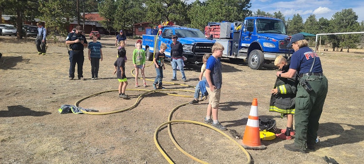 Outdoor event with firefighters demonstrating equipment to attendees, fire trucks in background, and hoses on the ground.