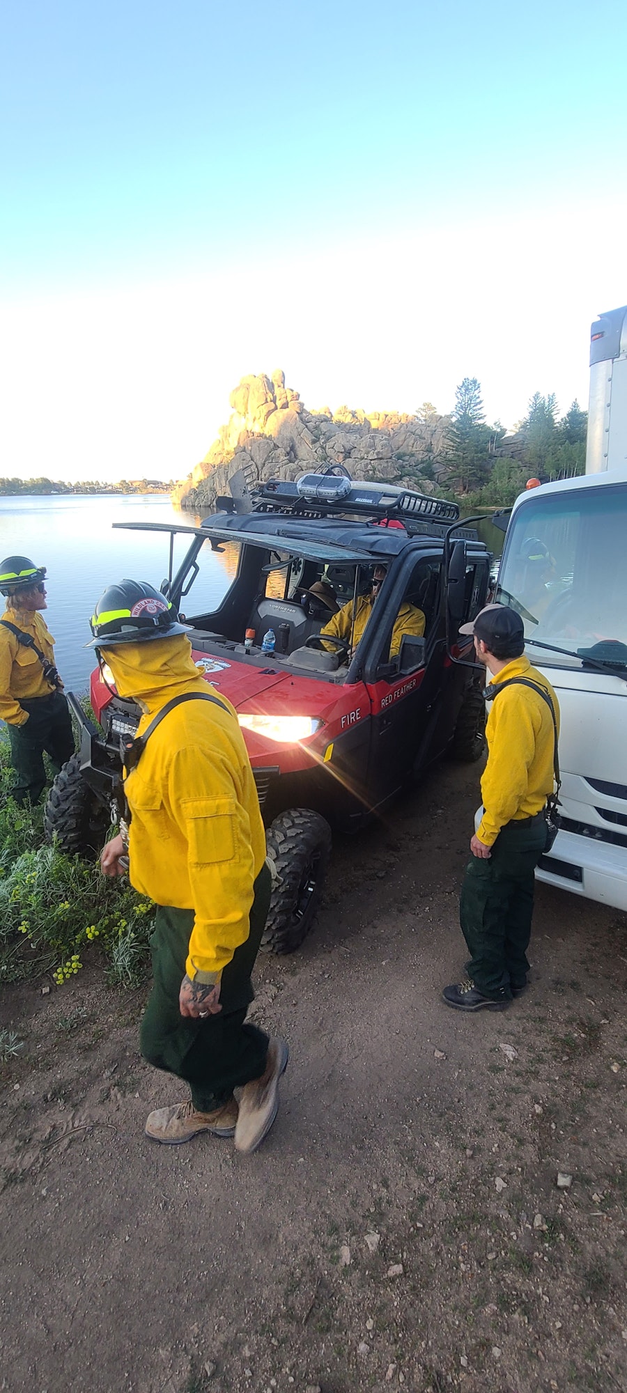People in yellow uniforms next to vehicles on a dirt path with rocky and forested scenery.