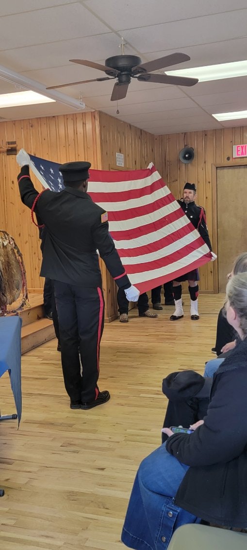 Two uniformed individuals holding an American flag indoors with an audience present.