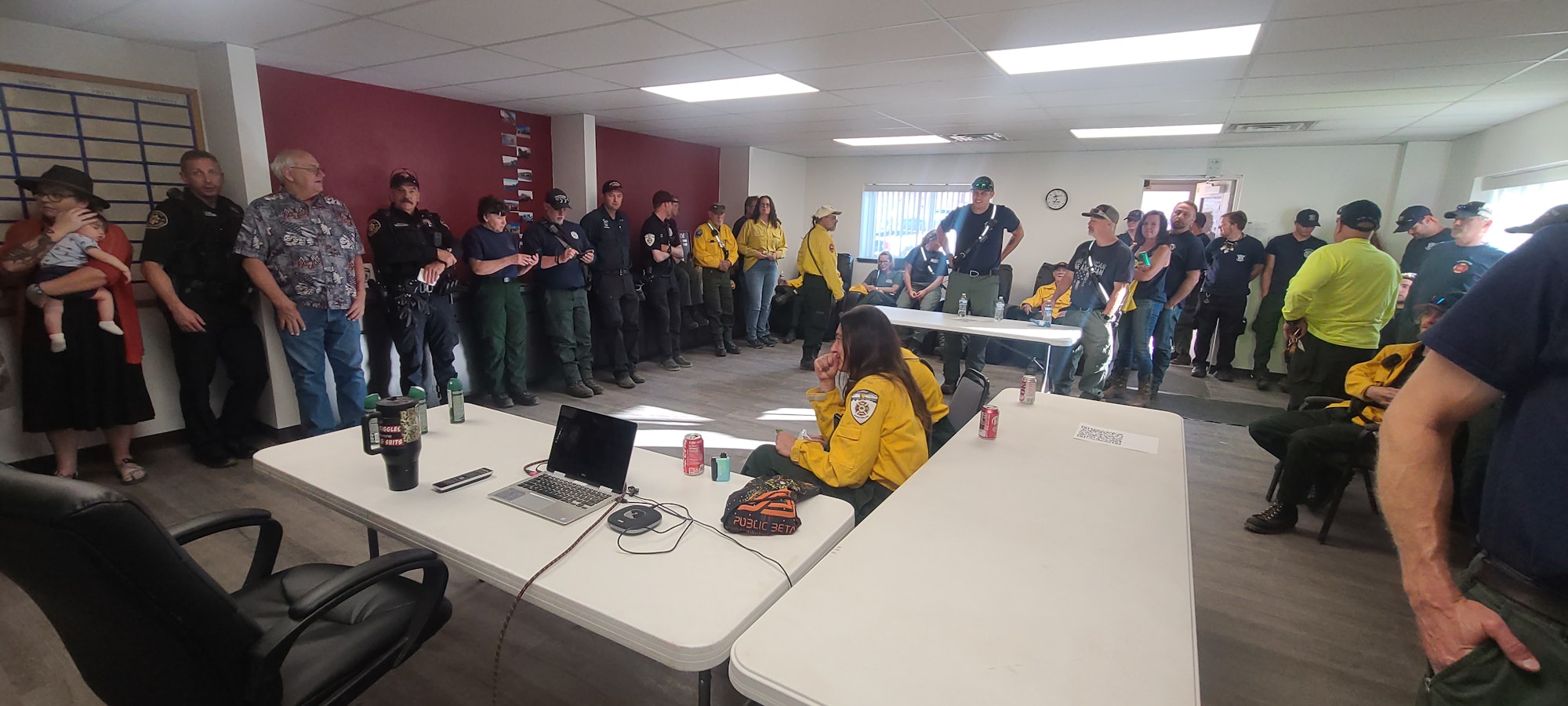 A group of emergency personnel in a meeting room, some in uniform, gathered around a table with laptops and drinks.