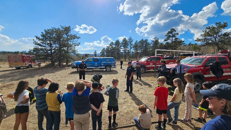 A group of children and adults attending a demonstration or event with emergency vehicles and personnel outdoors.