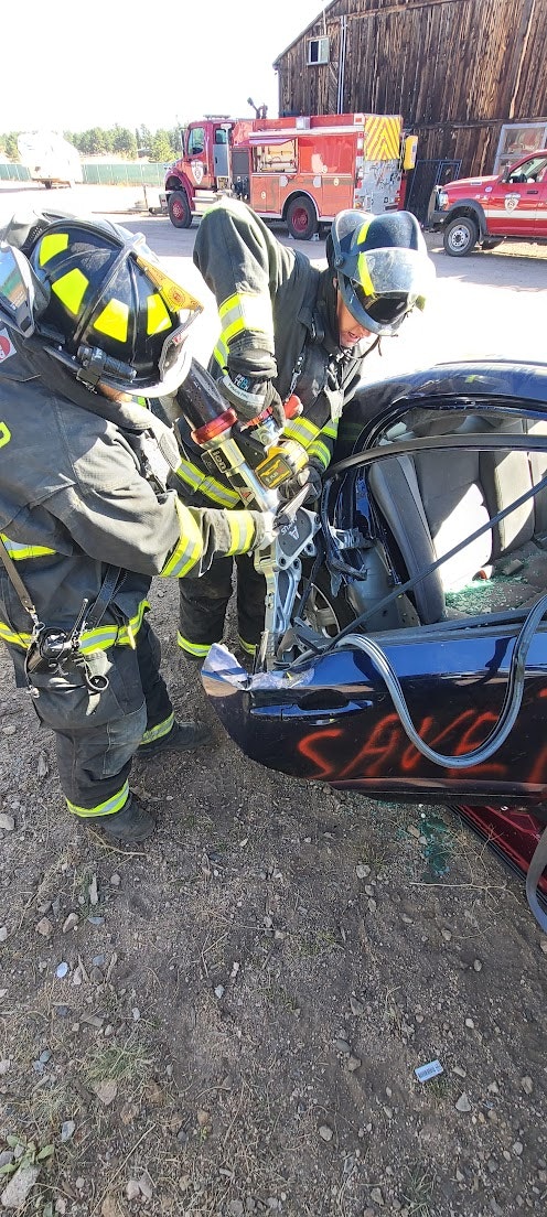 Firefighters use Jaws of Life to pry open car door; training or rescue scene with firetruck in background.