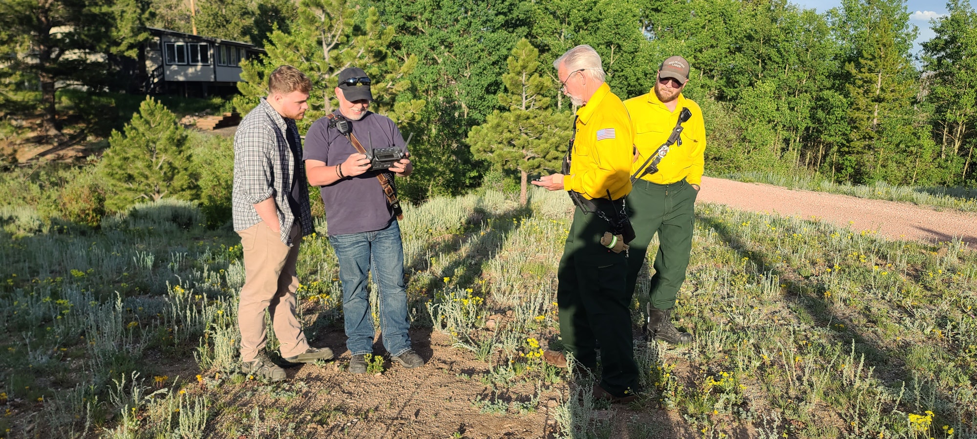 Four people standing outdoors in a grassy area, two in yellow shirts, two observing a device.