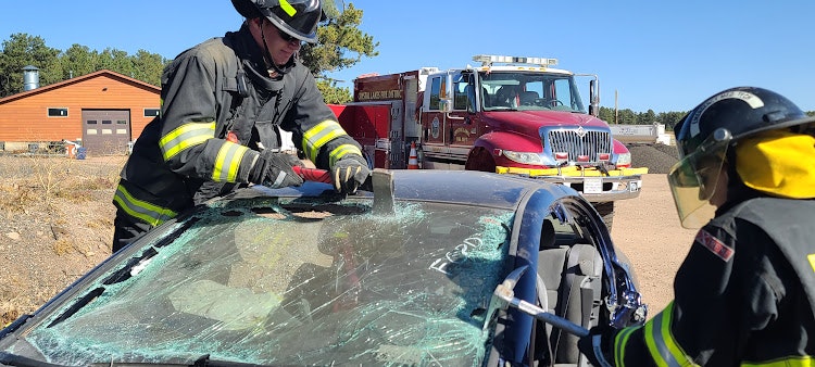 Firefighters practice vehicle extrication; damaged car, fire engine in background.