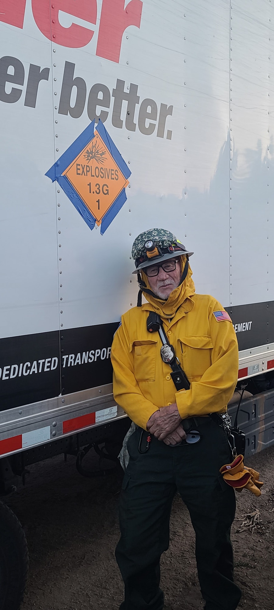A person in a yellow jacket and helmet stands by a truck with an "Explosives 1.3G" sign.