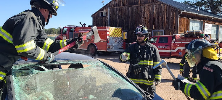 Firefighters training on car rescue with equipment, fire engine in background.