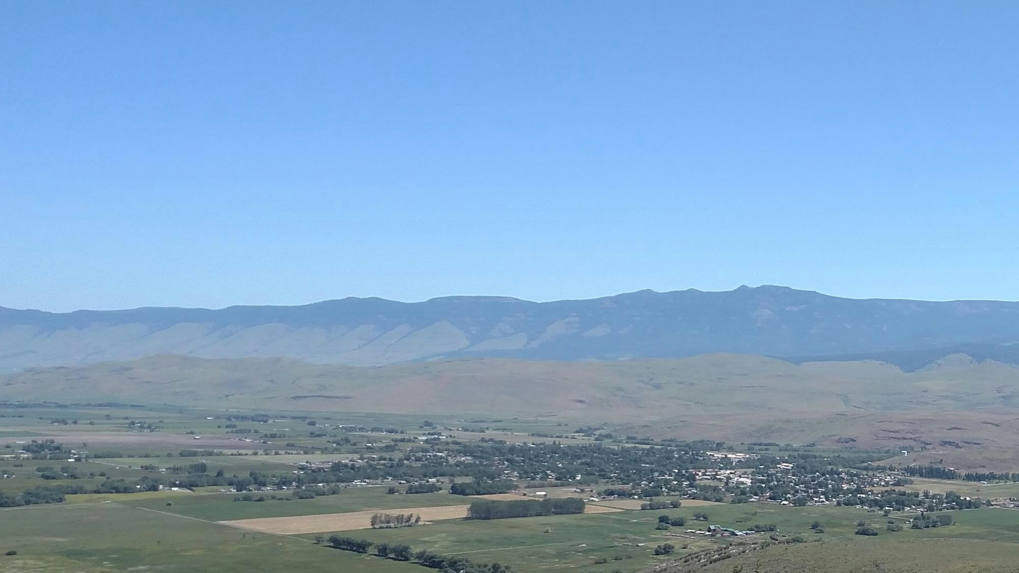 Rural landscape with a small town, fields, and a mountain backdrop under a clear sky.