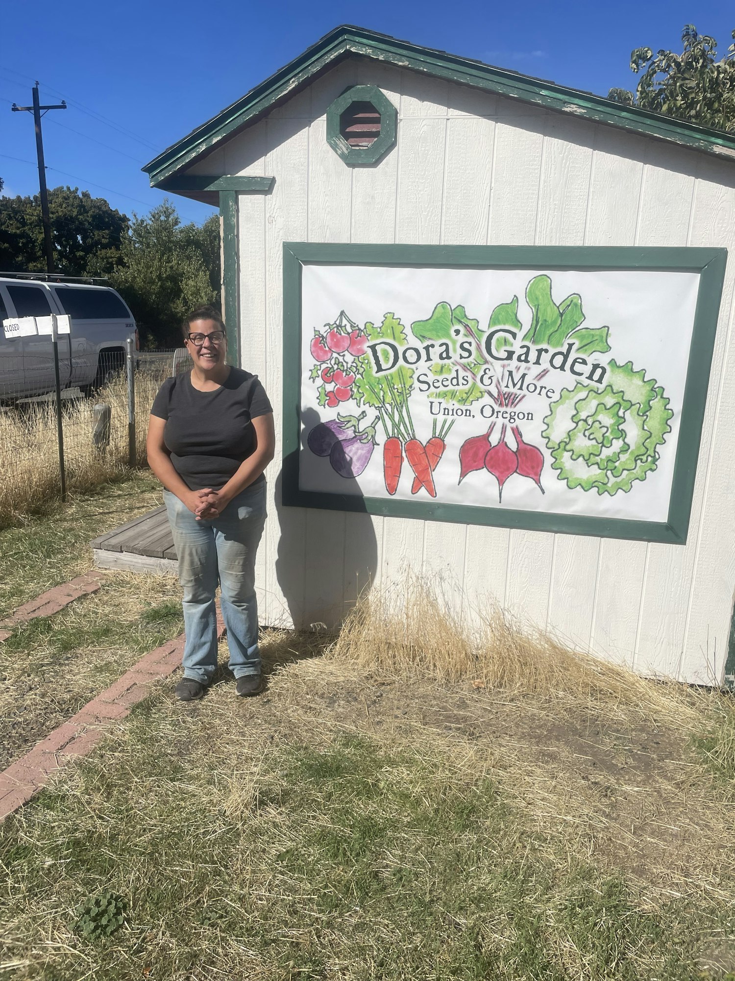 A person stands beside a sign for "Dora's Garden: Seeds & More" in Union, Oregon, on a sunny day.